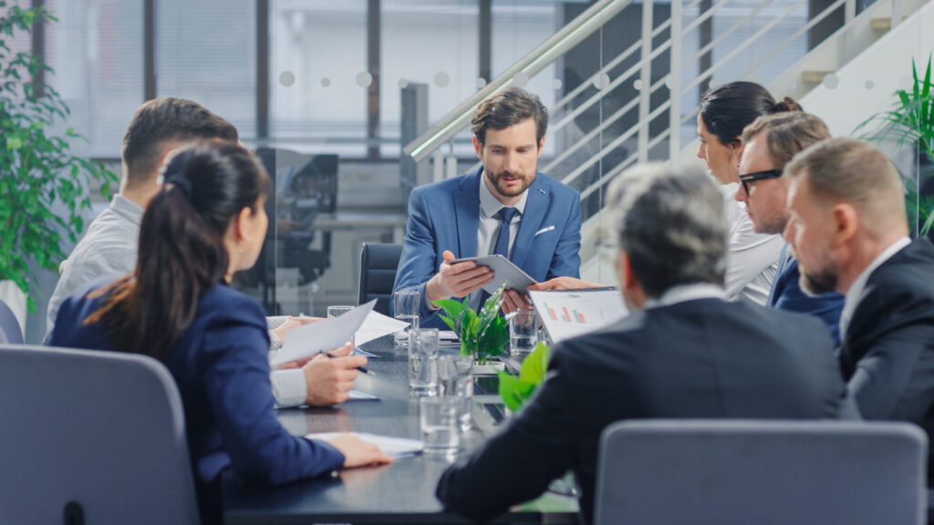In the Modern Corporate Office Meeting Room: Diverse Group of Businesspeople, Lawyers, Executives and Members of the Board of Directors Talking, Negotiating, Use Documents and Consult Statistic Graphs